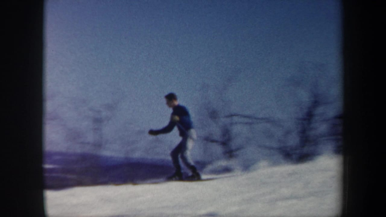 1962 Vermont 8mm frame showing a skier in a blue sweater and light pants crossing a snowy hillside with bare winter trees.
