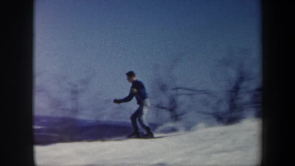 1962 Vermont 8mm frame showing a skier in a blue sweater and light pants crossing a snowy hillside with bare winter trees.