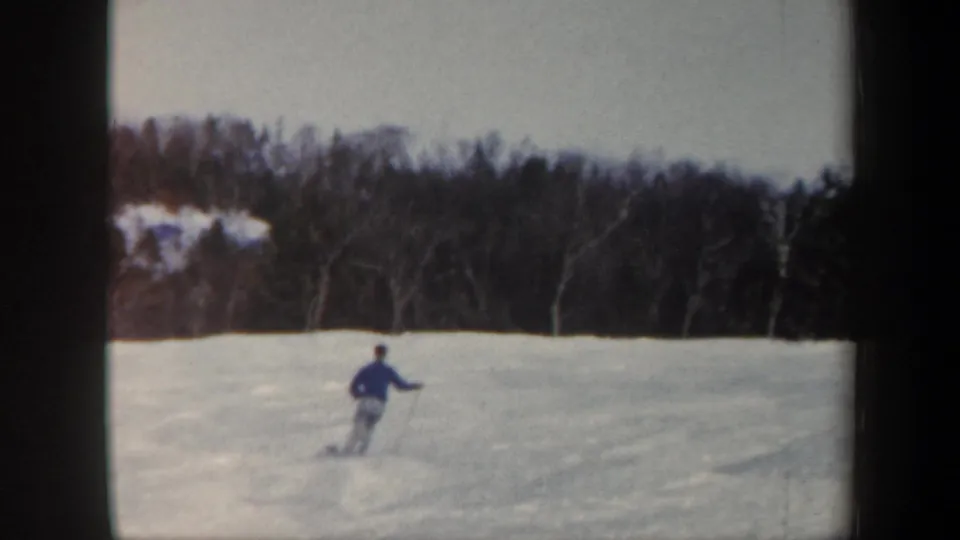 1962 Vermont 8mm frame showing a distant skier in blue moving across a snowy slope bordered by winter trees.