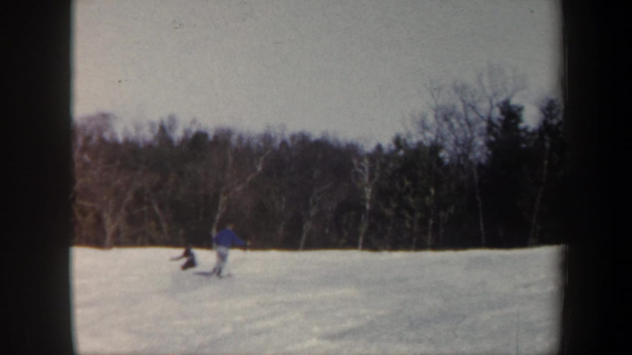 1962 Vermont home-movie frame with a skier in blue near another distant person on the snow, backed by winter woods.