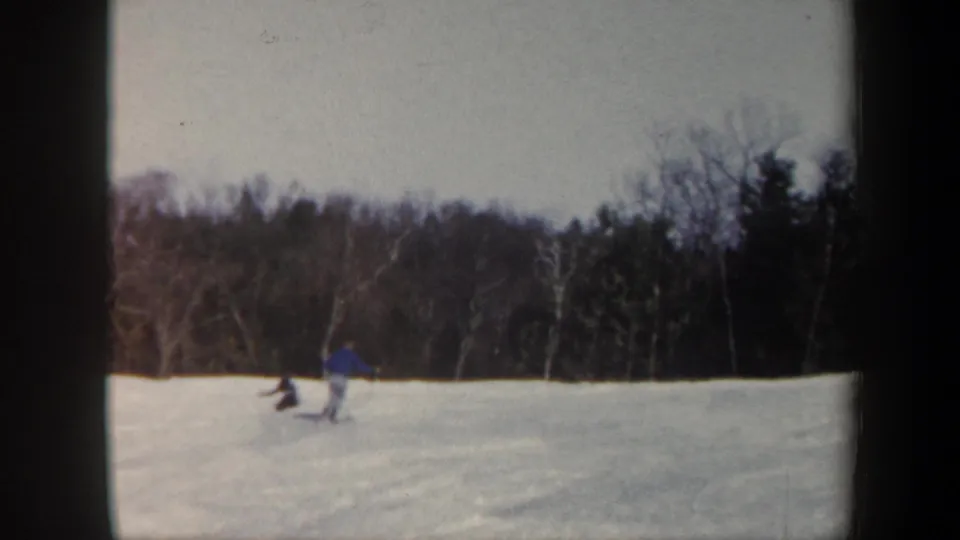 1962 Vermont home-movie frame with a skier in blue near another distant person on the snow, backed by winter woods.