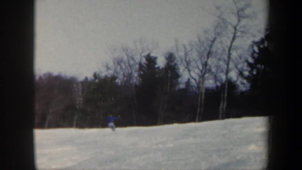 Distant 1962 Vermont skiing frame with the blue-clad skier lower on the slope beneath bare trees and evergreens.