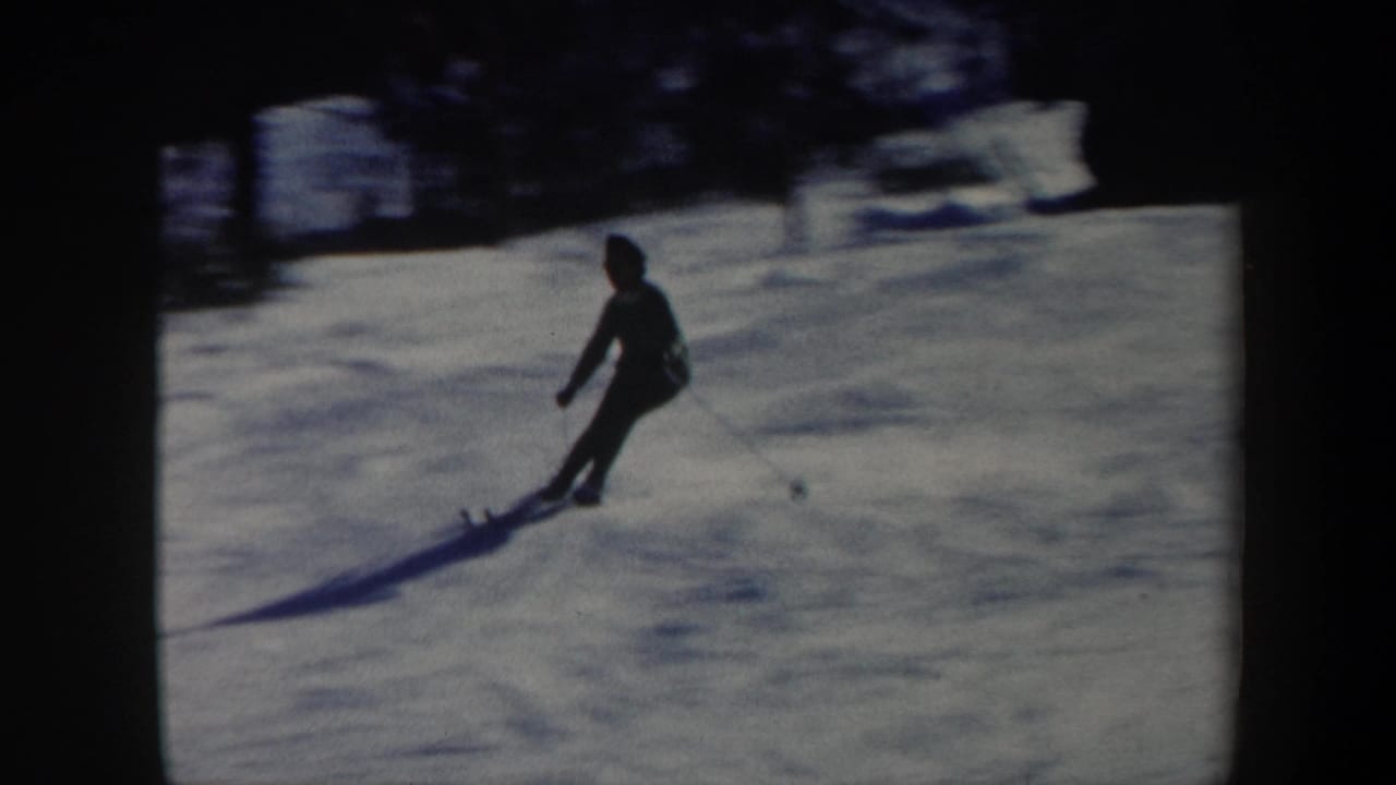In 1962 Vermont home-movie footage, a skier moves downhill across a snowy slope with poles, framed by dark trees and long shadows.