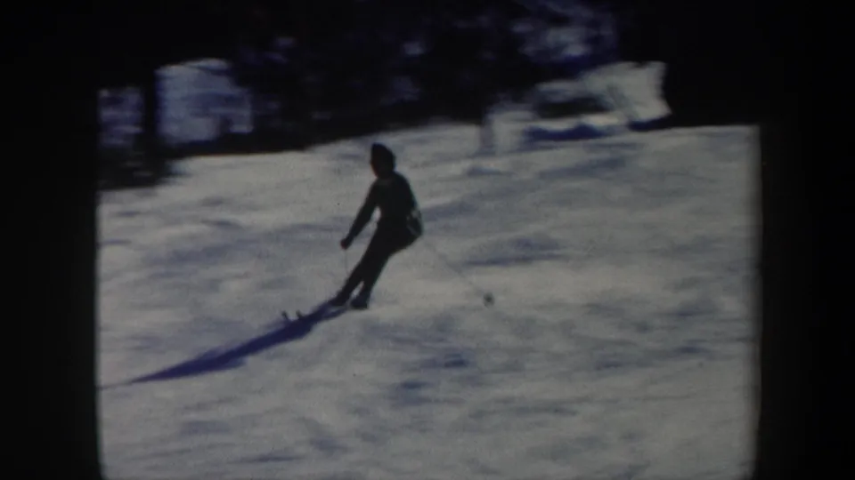 In 1962 Vermont home-movie footage, a skier moves downhill across a snowy slope with poles, framed by dark trees and long shadows.
