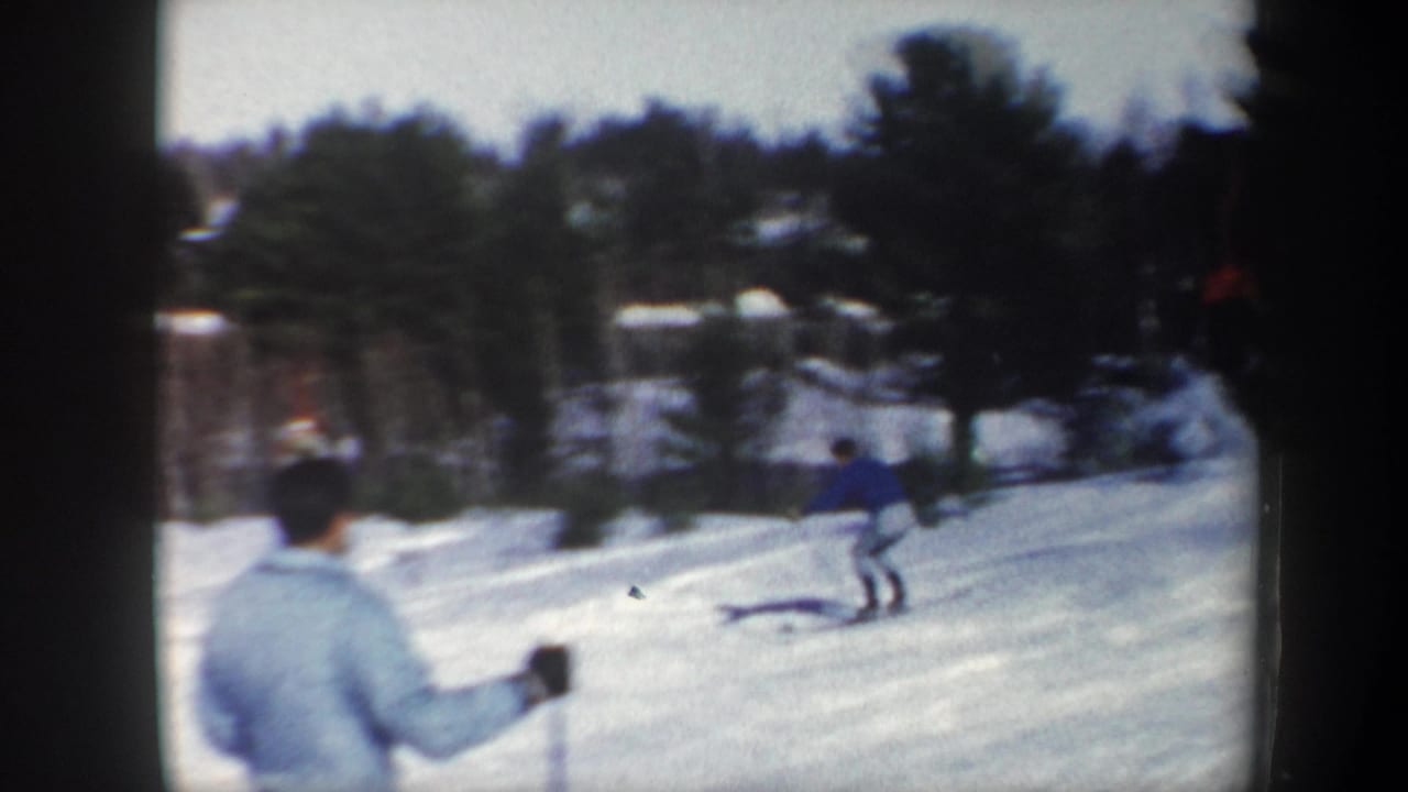 1962 Vermont home-movie frame showing a skier practicing on snow while a foreground figure watches near evergreen trees.