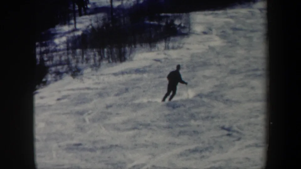 1962 Vermont frame of a skier turning downhill on a broad snowy slope with winter brush and trees at the edge.