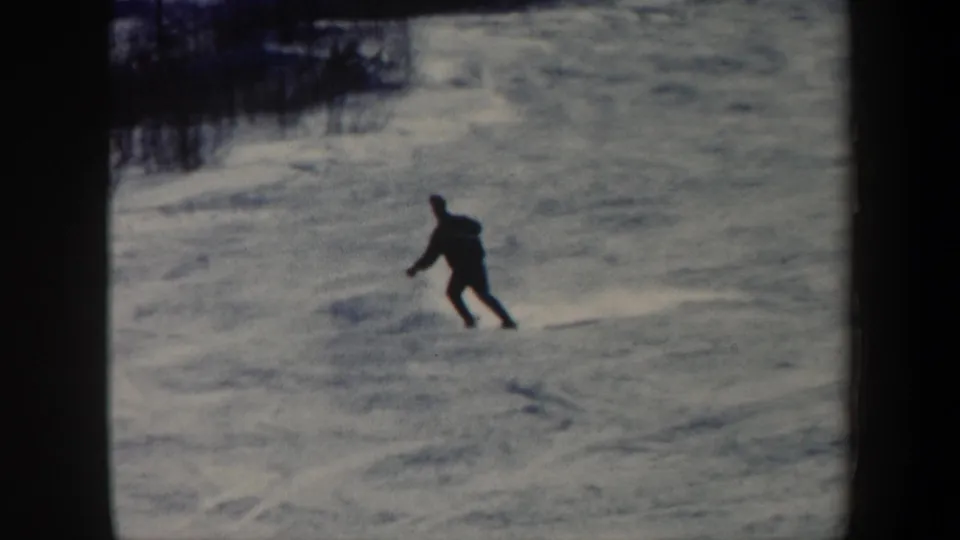 A skier in 1962 Vermont leans into a turn across a textured snow surface, seen in soft 8mm home-movie footage.