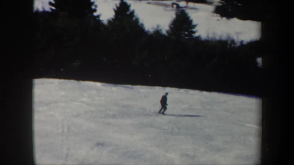 Small figure of a skier crossing a snow-covered Vermont slope, framed by evergreens in archival 8mm footage.