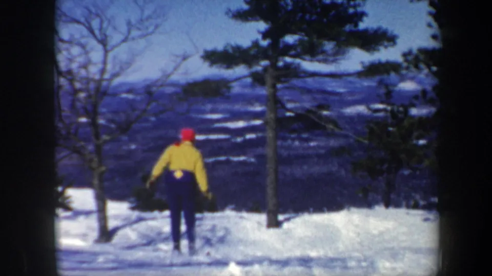 1962 Vermont frame showing a skier in a yellow jacket and red hat moving away across snow between trees.