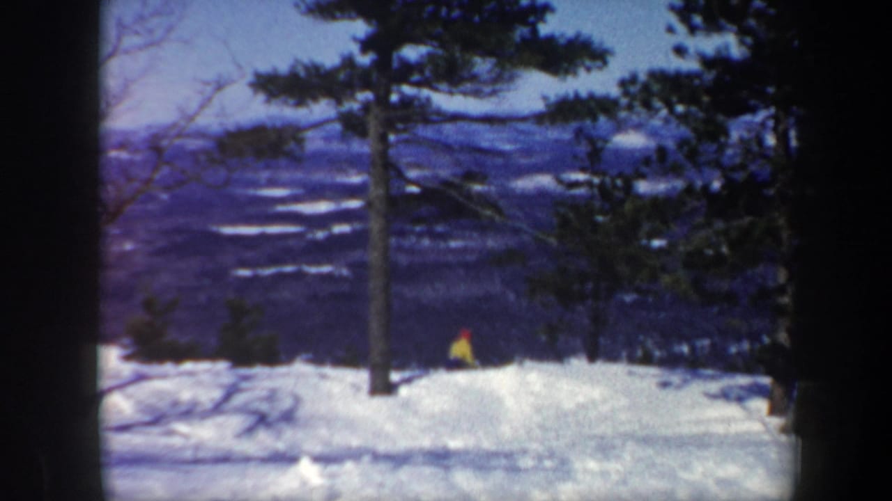 Distant skier in bright clothing moves across a snowy rise near trees.