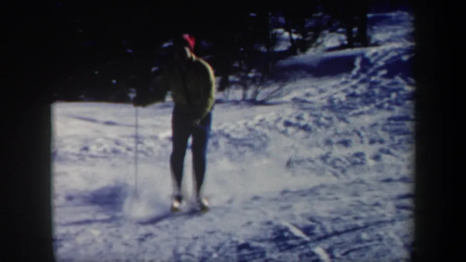 1962 Vermont home-movie frame showing a skier in a yellow jacket and red hat on a tracked snowy slope.