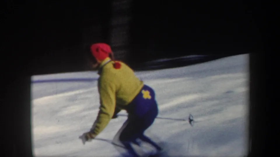 Close 1962 Vermont frame of a skier in a yellow jacket and red hat bending into a downhill turn on packed snow.