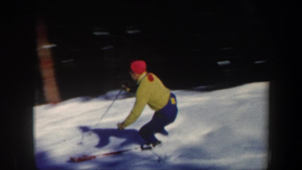 1962 Vermont home-movie frame showing a skier in a bright yellow jacket and red hat carving through powder on a shaded slope.