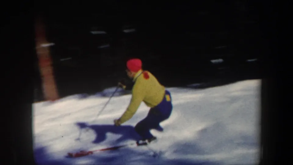 1962 Vermont home-movie frame showing a skier in a bright yellow jacket and red hat carving through powder on a shaded slope.