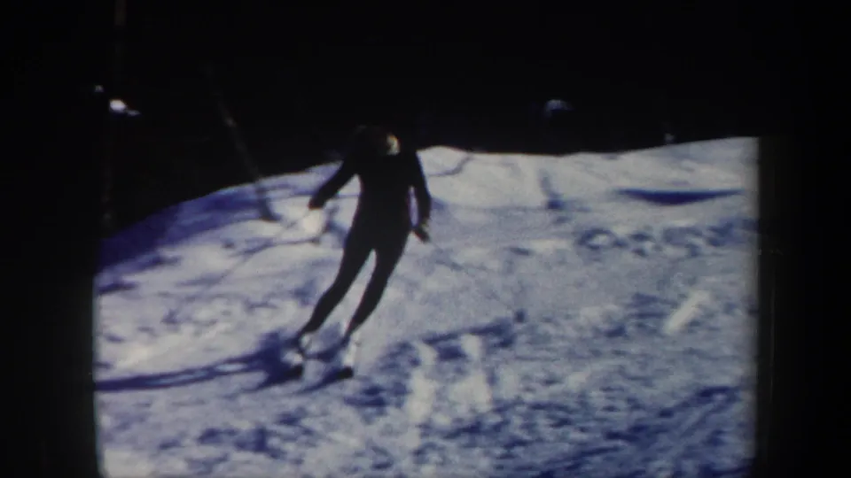 A skier in dark winter gear carves across a powder-covered Vermont slope in grainy 8mm home-movie footage.