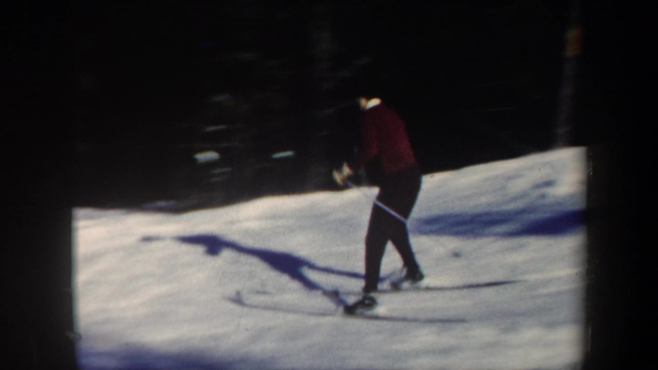 Skier in red crosses a bright snowy slope with a long shadow on the snow.