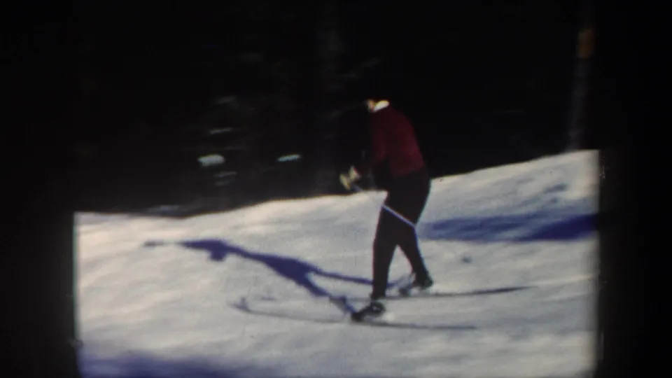 1962 Vermont ski footage frame with a red-clad skier using poles on a sunlit snow slope beside dark evergreens.
