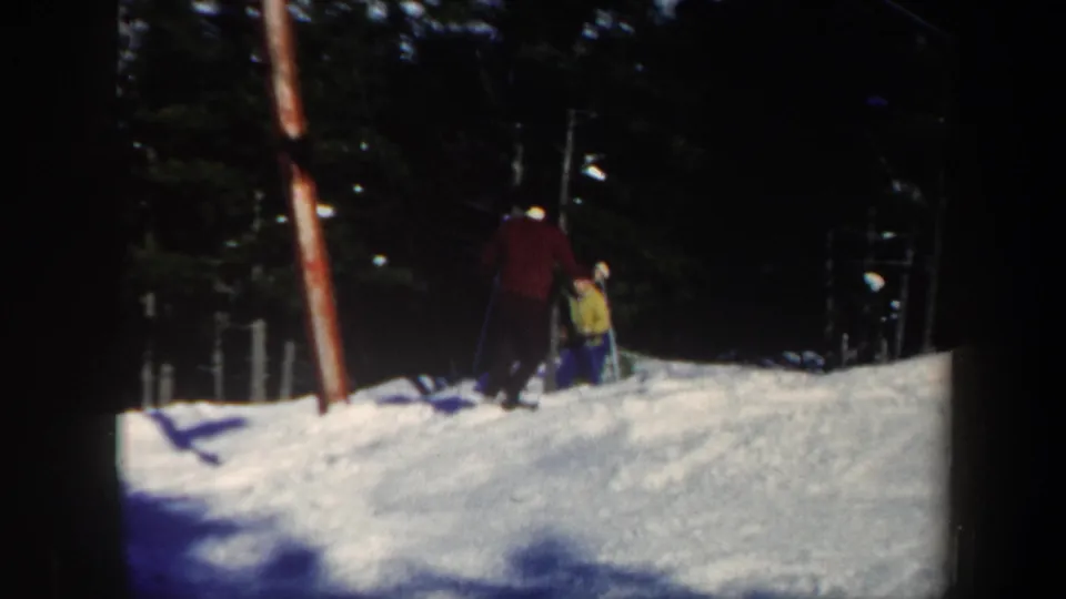 Home-movie frame from Vermont in 1962 showing a skier in red near another person in yellow on a snowy slope by trees.