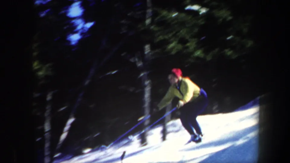 1962 Vermont home-movie frame with a skier in a yellow jacket and red hat moving downhill through trees.