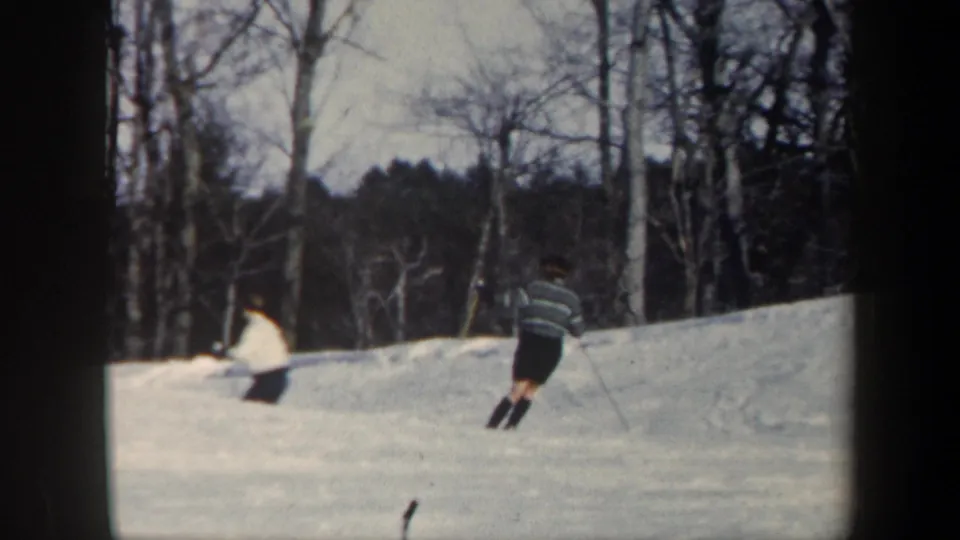 1962 Vermont home-movie frame showing two skiers on a snowy hillside bordered by leafless trees.