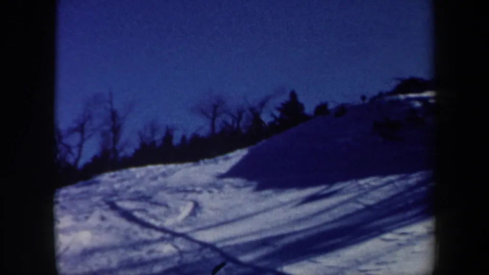 Blue-tinted 8mm frame of a snowy Vermont ski slope with curved tracks and a dark wooded ridge.