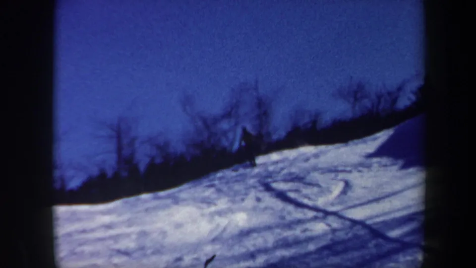 Blue-tinted home-movie frame showing a distant skier on a snowy Vermont slope with tracks and bare trees.