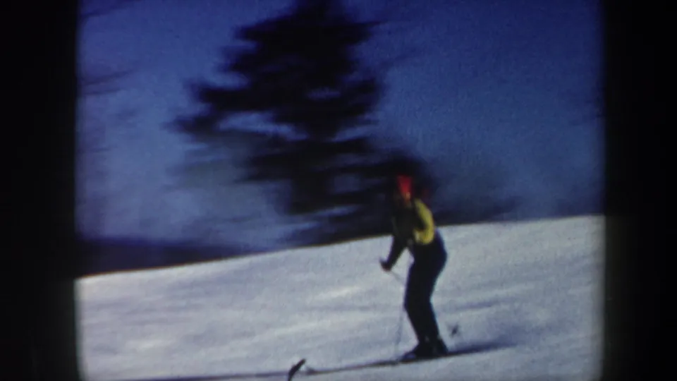1962 Vermont 8mm frame showing a blurred skier in yellow and red moving quickly past a snowy slope and dark evergreen.