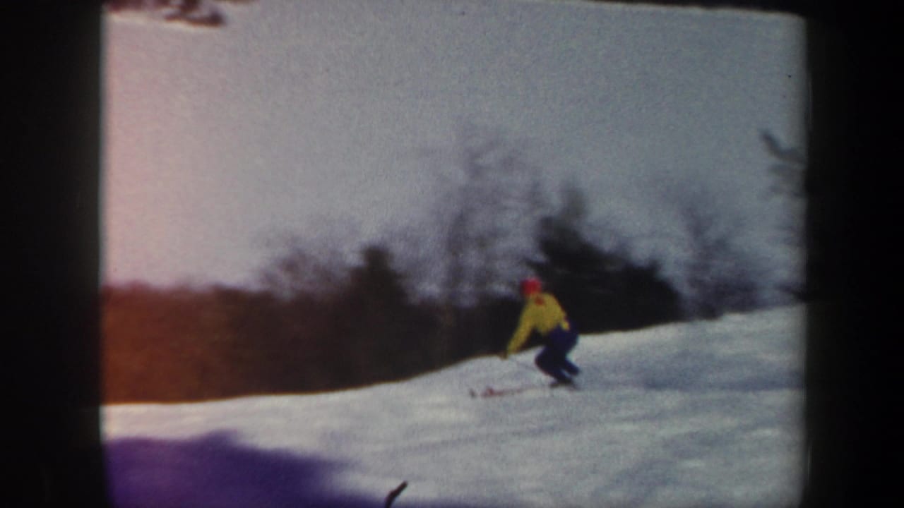 A skier in a yellow jacket and red hat moves downhill on a snowy Vermont slope in 1962 home-movie footage, with trees lining the background.
