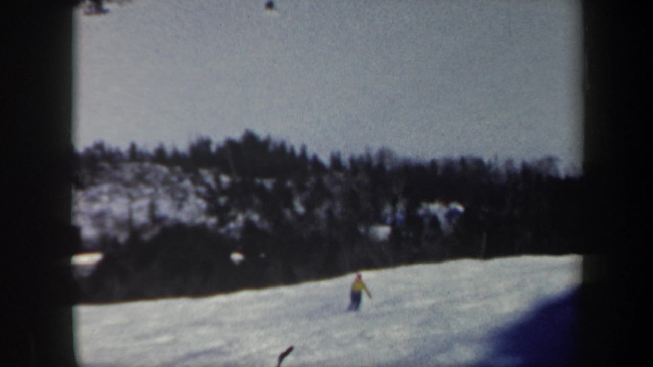 Skier continues downhill on a snowy slope below a wooded ridge.