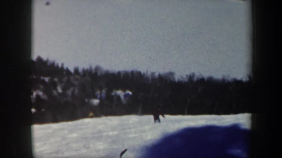 1962 Vermont 8mm frame showing the skier farther along the snowy slope, with dark woods and a blue foreground shadow.