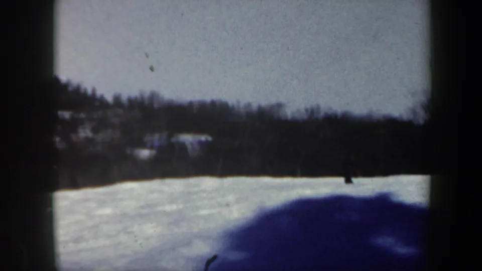 1962 Vermont home-movie frame with the skier near the right edge of the snowfield under a dark winter treeline.