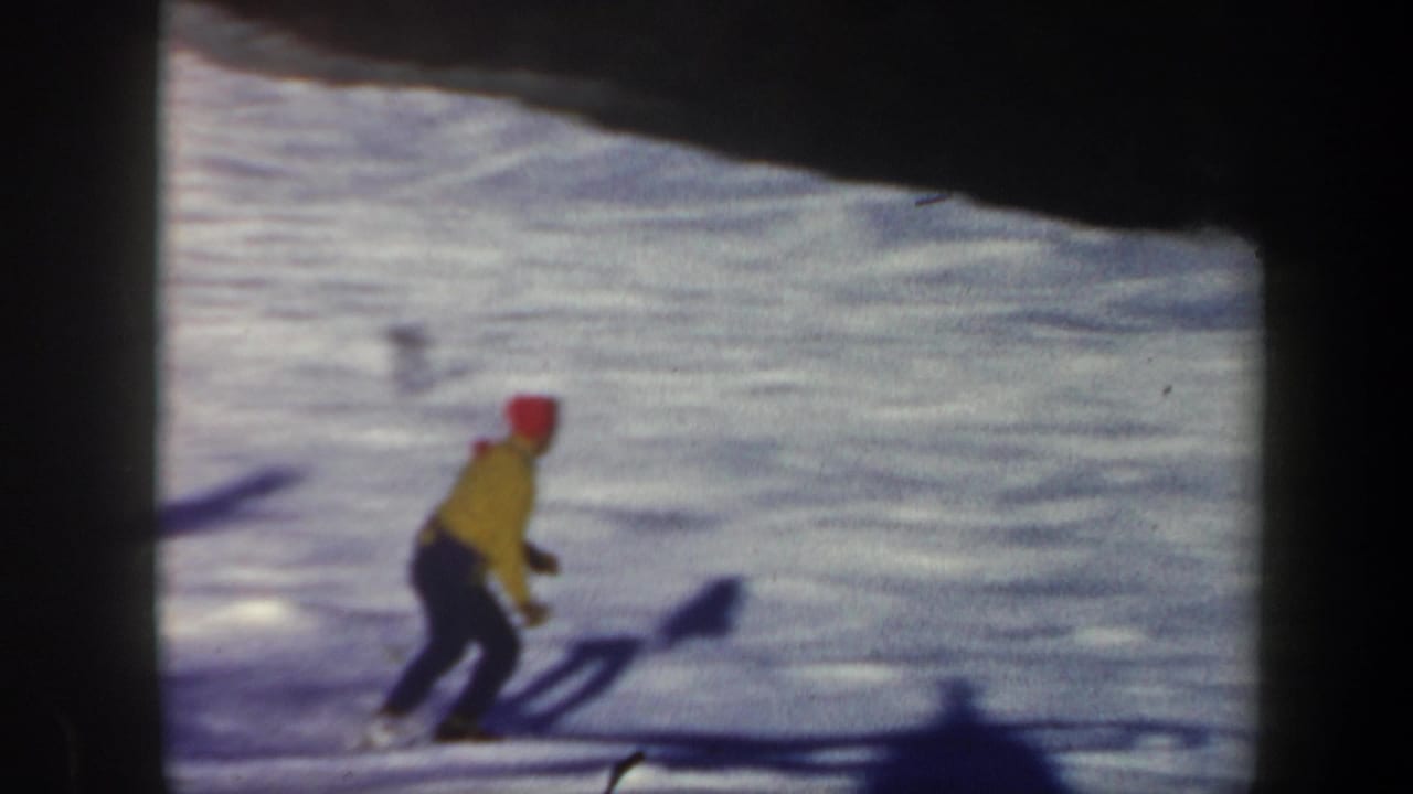 1962 Vermont 8mm frame showing a lone skier in a yellow jacket and red hat on a bright snowy slope beside dark woods.