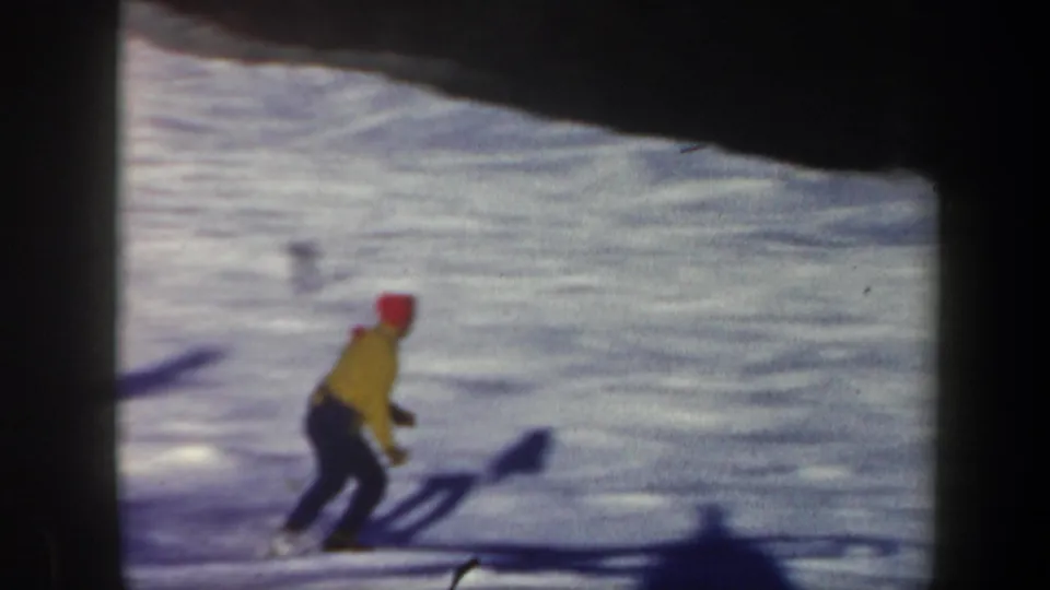 1962 Vermont 8mm frame showing a lone skier in a yellow jacket and red hat on a bright snowy slope beside dark woods.