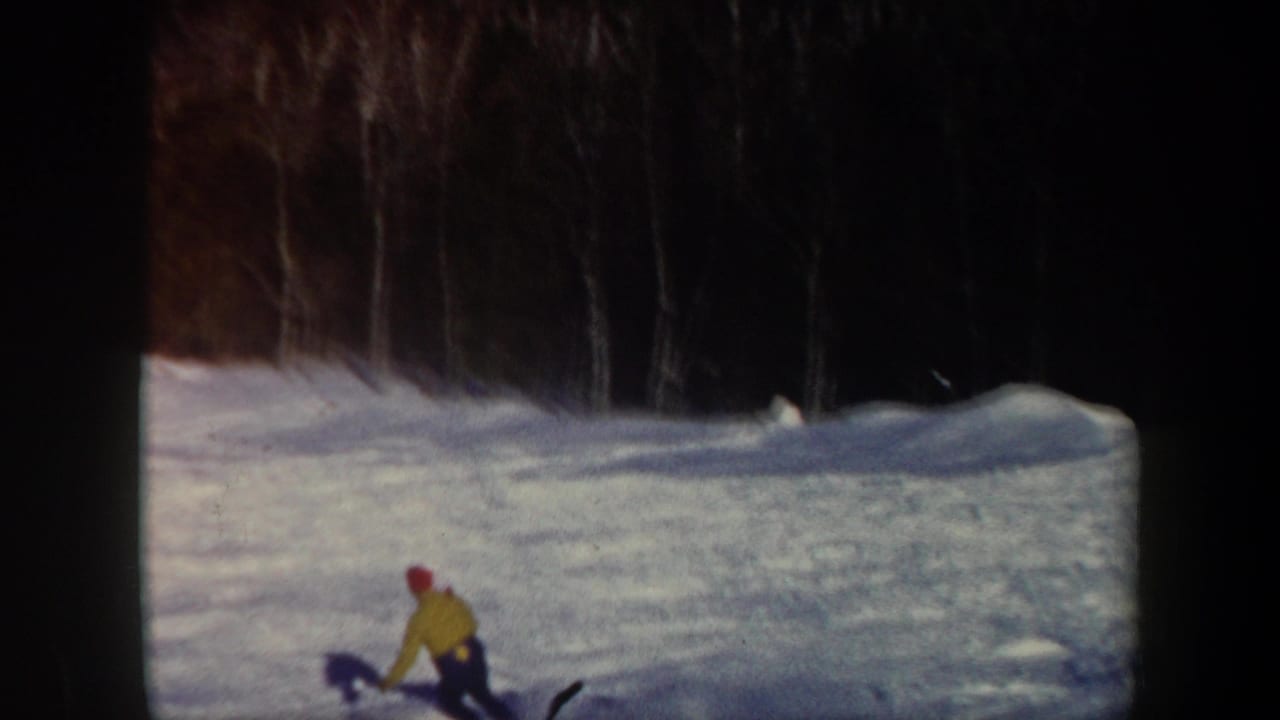 1962 Vermont home-movie frame with a distant skier in yellow descending a white slope below a dark winter tree line.
