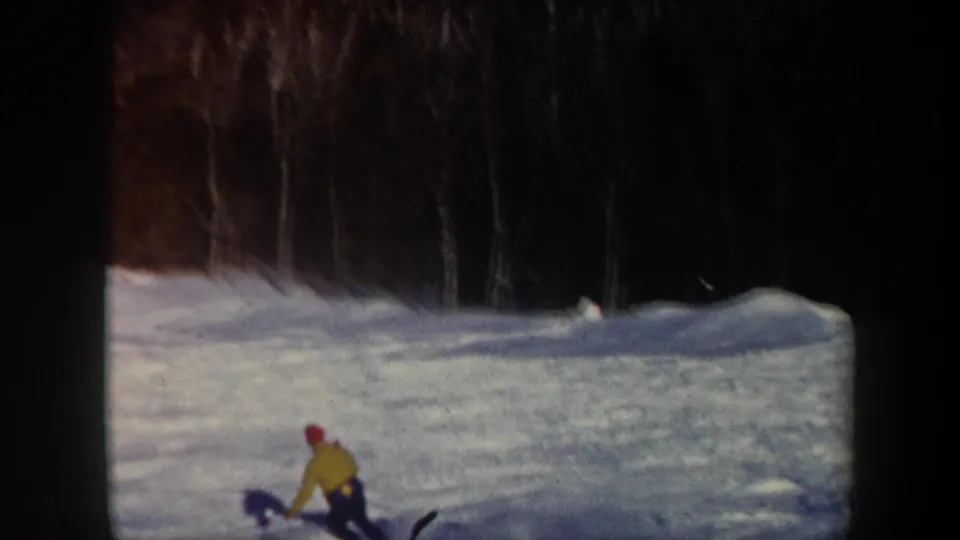1962 Vermont home-movie frame with a distant skier in yellow descending a white slope below a dark winter tree line.