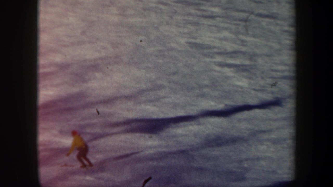 A distant skier in a yellow jacket crosses a snowy slope with long shadows.