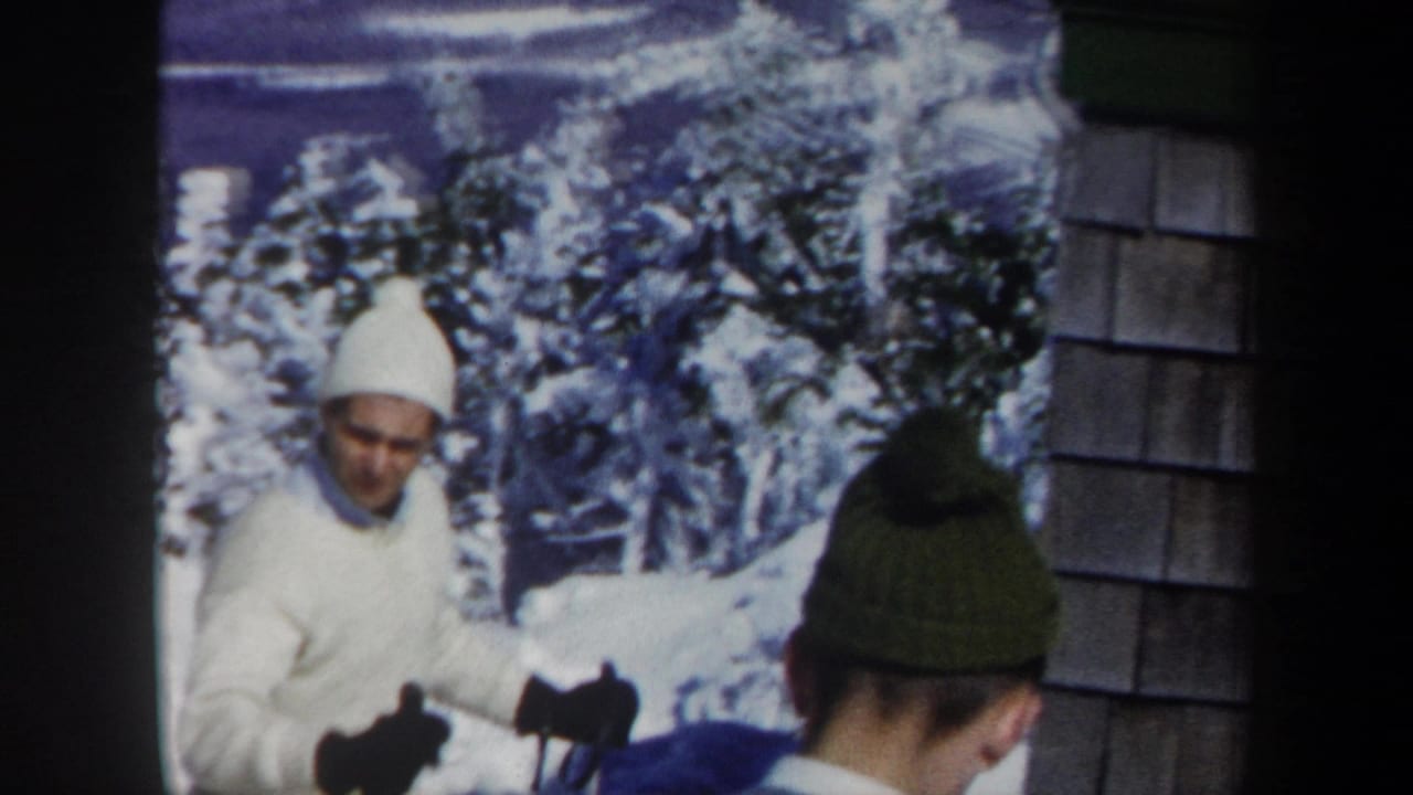 1962 Vermont home-movie frame showing a man in white ski clothing near a shingled building, with another skier seen from behind.