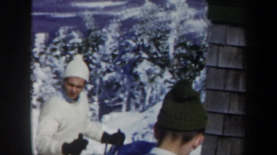 1962 Vermont home-movie frame showing a man in white ski clothing near a shingled building, with another skier seen from behind.