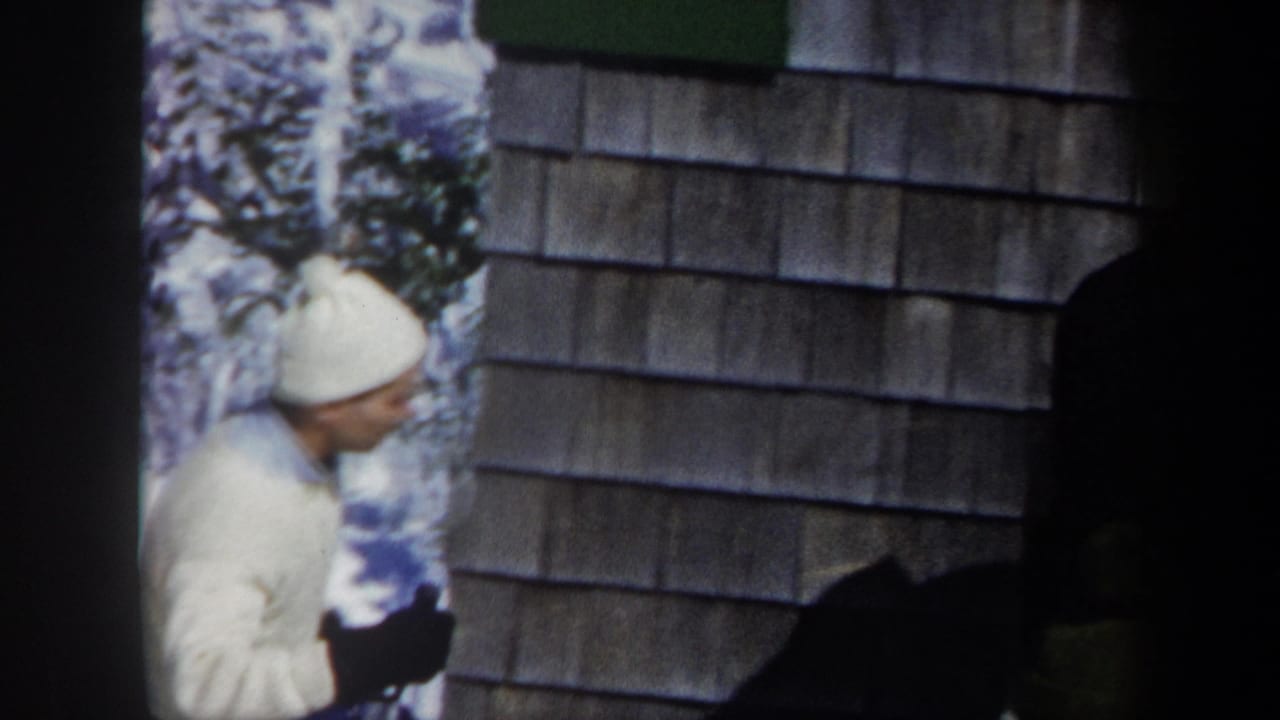 Man in a white hat and ski clothing stands beside a wood-shingled wall in snow.