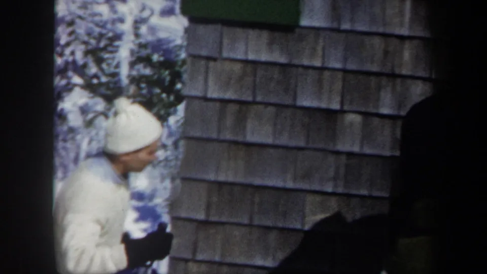 1962 Vermont frame with an adult skier in white hat and clothing beside a shingled building, holding dark gloves or gear.