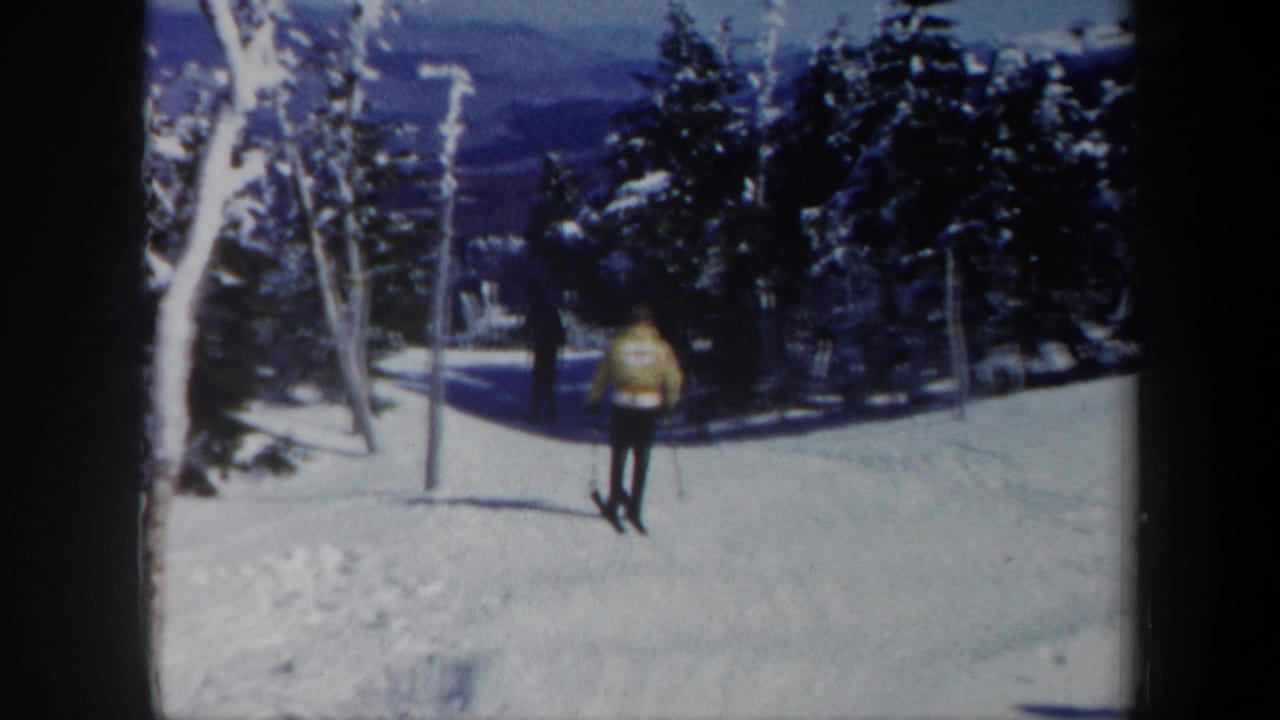 A skier in a yellow jacket approaches on snow while another skier follows behind.