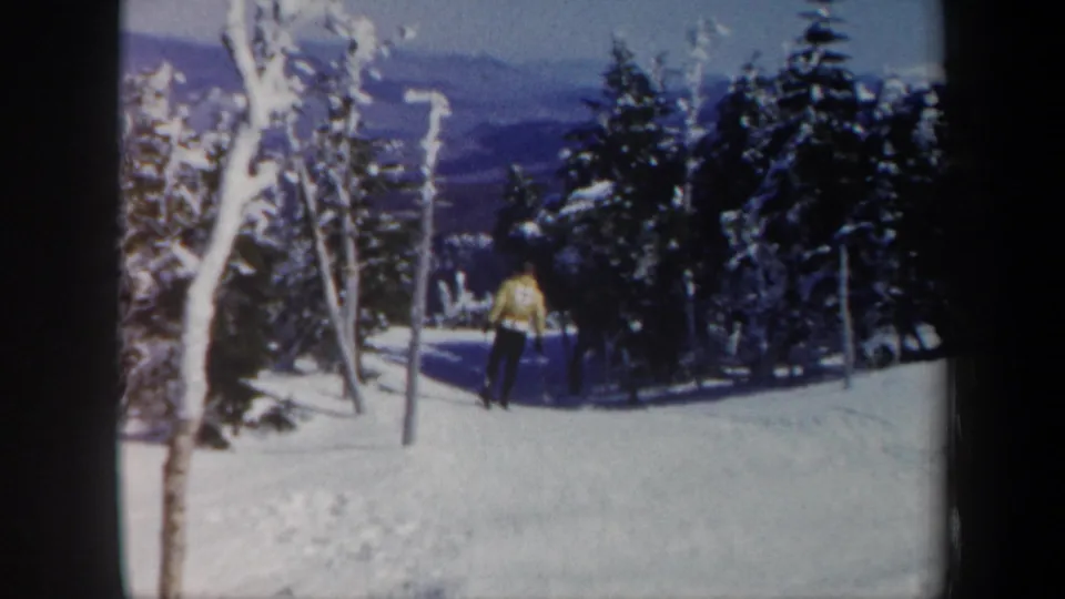 1962 Vermont home-movie frame showing the foreground skier continuing down the snow-covered run between trees.