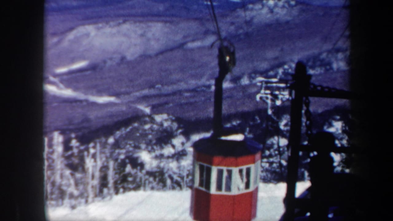 Red gondola near dark cableway equipment with snow and valley scenery behind it.