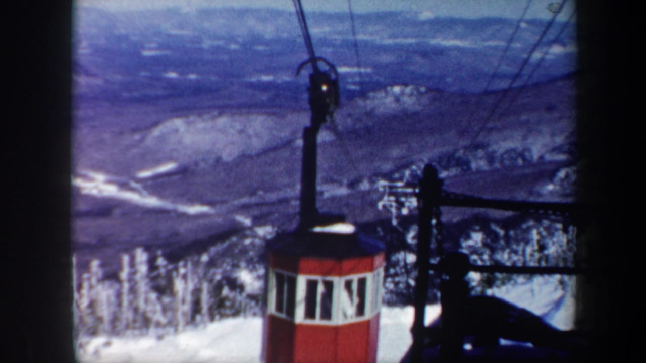 1962 Vermont home-movie frame showing a red gondola cabin on overhead cables with snowy forested slopes and distant ridges behind it.