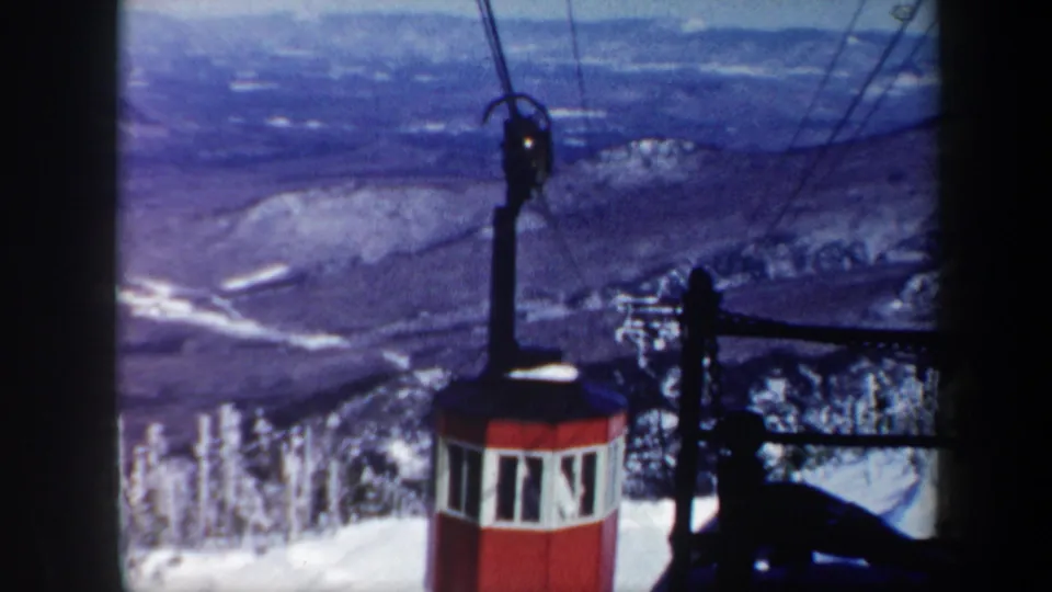 1962 Vermont home-movie frame showing a red gondola cabin on overhead cables with snowy forested slopes and distant ridges behind it.