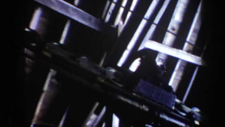 1962 Vermont home-movie frame showing a close, shadowed view of ski lift machinery with rails, wheels, and reflective metal rods.