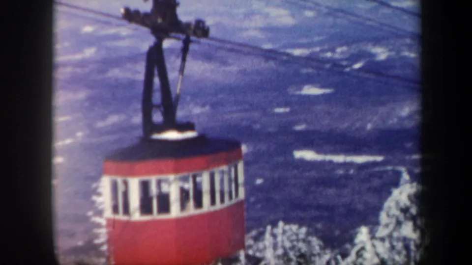 1962 Vermont frame showing a red aerial cable car moving above snowy trees and distant mountains.
