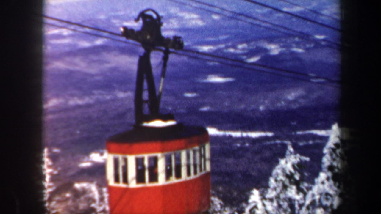 1962 Vermont 8mm frame showing a red and white cable car above snow-covered evergreens with distant winter terrain behind it.