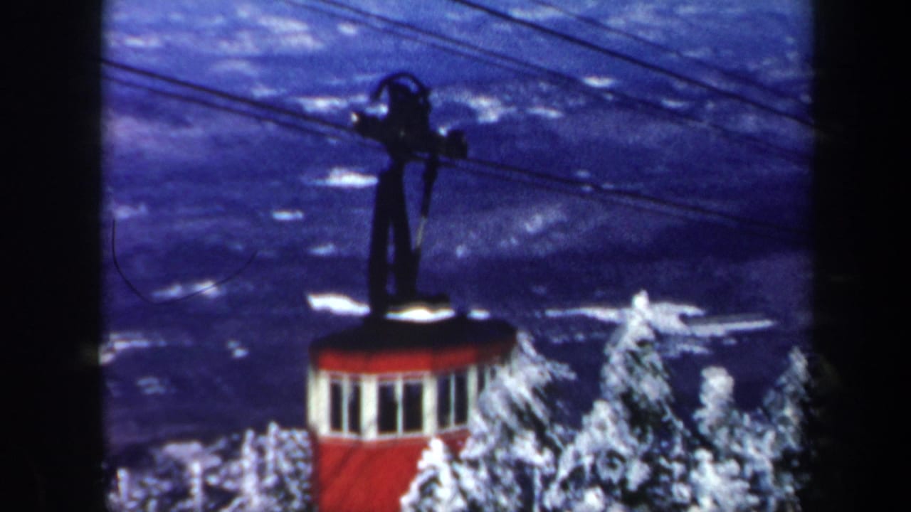 1962 Vermont 8mm frame with the red tram cabin lower in view, snowy trees at right, and cable lines stretching across a blue landscape.