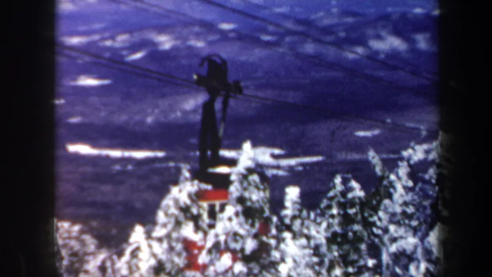 1962 Vermont 8mm frame showing the cable car farther away, partially obscured by snowy trees, with dark cables crossing the winter valley view.